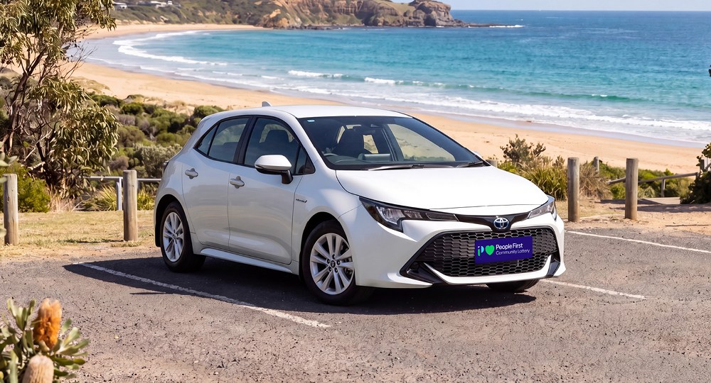 White Toyota Corolla hatchback parked at a coastal lookout overlooking a beach and ocean, featuring People First Community Lottery branding on the licence plate.
