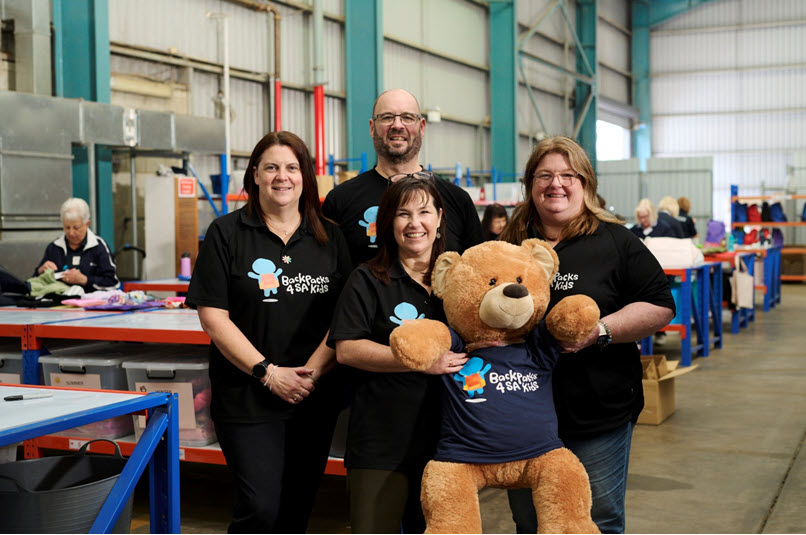 Four people wearing matching branded Backpacks for Kids shirts pose in a warehouse or workshop setting with a large teddy bear seated in front of them.