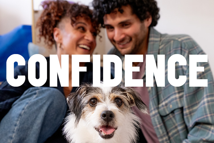 Two people sitting close together behind a small dog in a cardboard box indoors, with the word “Confidence” overlaid.