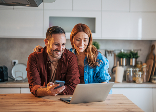Couple at kitchen bench smiling at laptop.