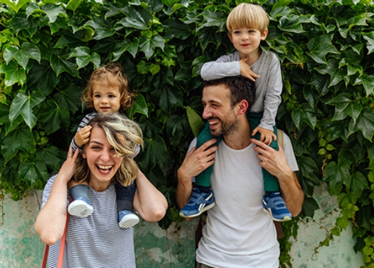 A couple carry their kids on their shoulders in front of a vine covered wall.