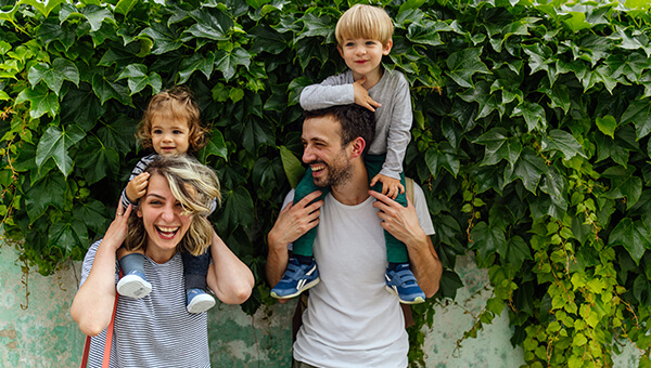 A couple carry their kids on their shoulders in front of a vine covered wall.