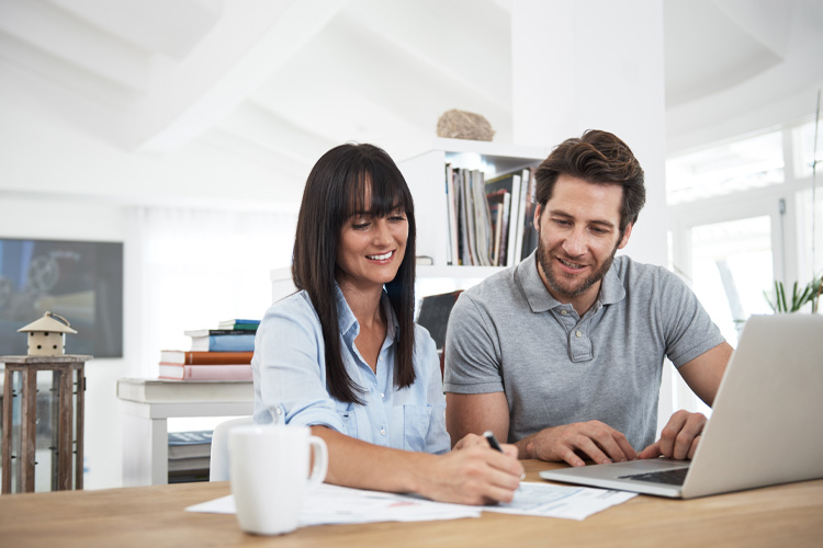 Two people at table, one writing on paper, the other using a laptop, with bookshelves in background.