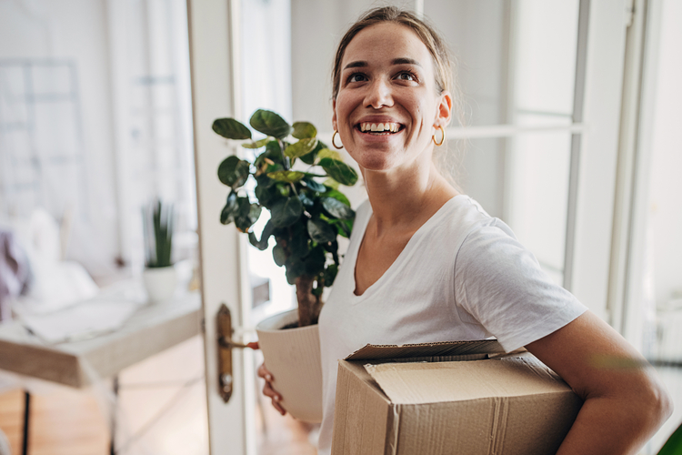 Woman carrying plant and boxes into new home.