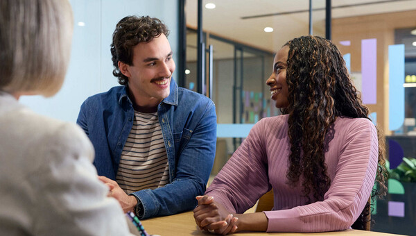 A People First Bank employee talking to two customers seated at a table inside a branch. 