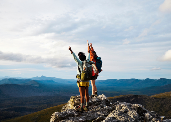 Two women with backpacks with their hands up, overlooking a mountain range.