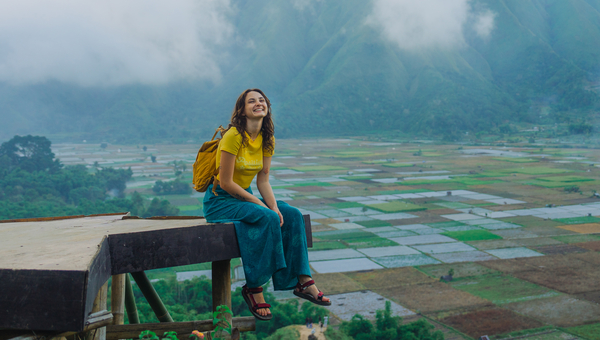 Young woman sitting on a wooden platform looking over farmland at the bottom of a mountain valley.