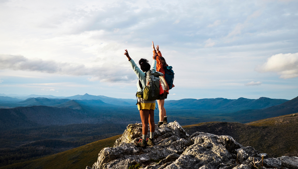 Two women with backpacks with their hands up, overlooking a mountain range.