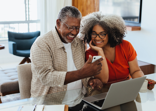 A grey-haired couple using a laptop while the man holds a phone.