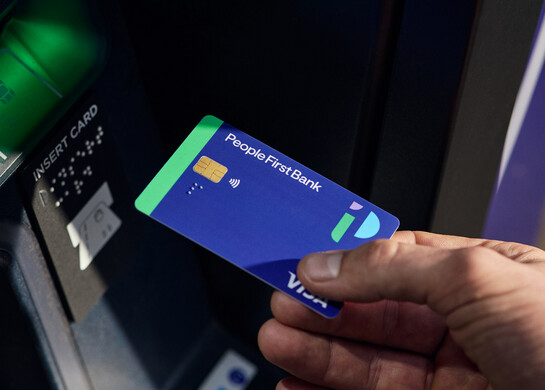 A hand inserting a People First Bank Visa debit card into an ATM. 