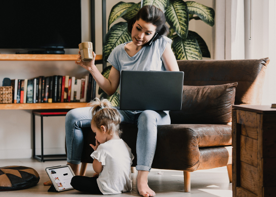 A mother sits with her laptop and her toddler sits on the floor with a tablet.