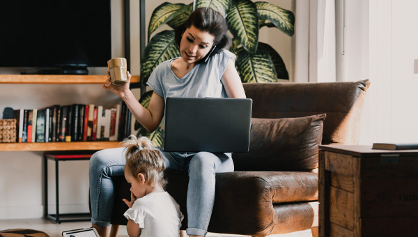 A mother sits with her laptop and her toddler sits on the floor with a tablet.