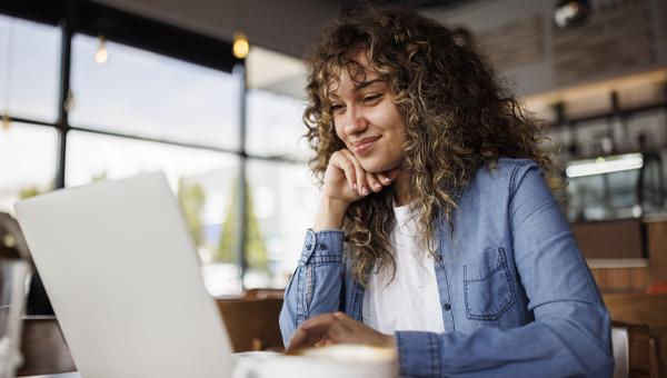 Woman sits at a table with a coffee in her kitchen, in front of her laptop.