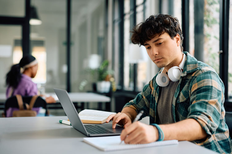 A man studies at his laptop. He has headphones around his neck.