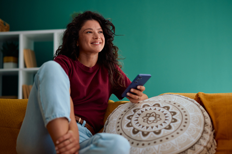 Woman sits on her couch with a cushion holding her phone.