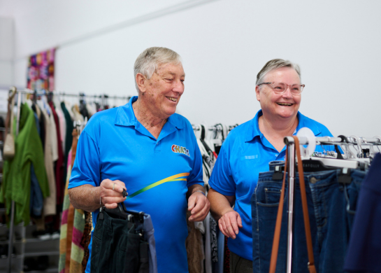 Two smiling volunteers in blue shirts browse donated clothing on racks in a community op shop.