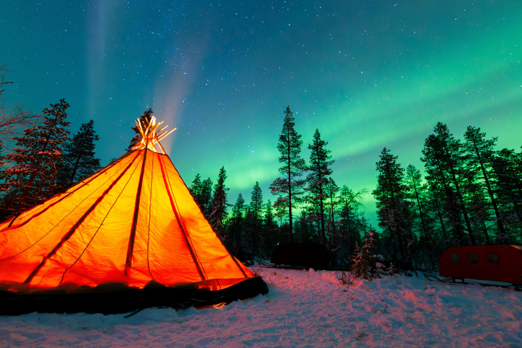 A tent on a snowy hill framed by trees glows from within, under a sky streaked with green lights.