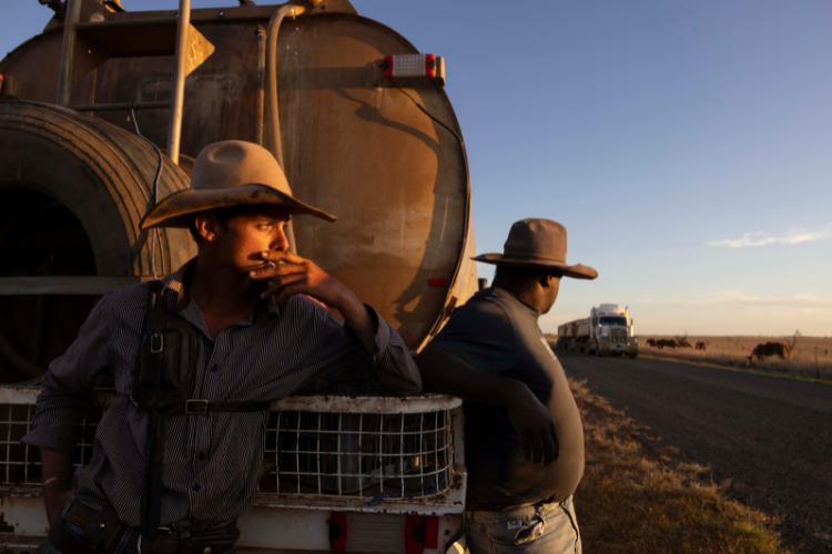 Two men in cowboy hats lean against the back of a water truck parked on an outback road, lit by warm sunlight.